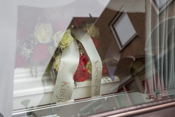 Een kist met linten en bloemen gezien door het raam van een rouwauto, afscheidsfotografie